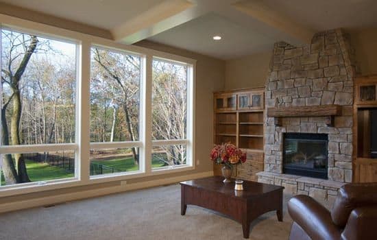A tan living room with a large stone fireplace and floor-to-ceiling picture and awning windows overlooking a yard.